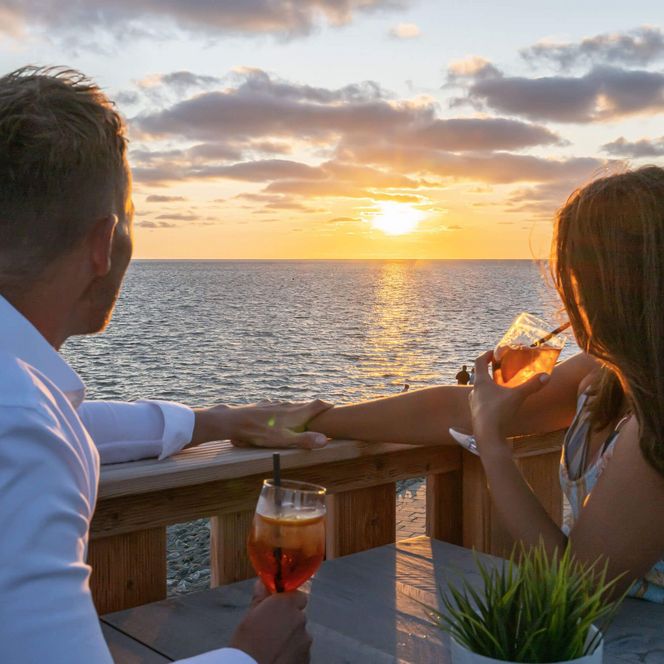 Ein junges Paar sitzt mit Cocktails auf der Dachterrasse der 360 Grad Bar in Büsum und betrachtet den Sonnenuntergang.