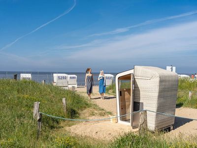 buesum-strandkorbsauna-freundinnen05 Zwei Frauen gehen, in Handtücher gekleidet zur geöffneten Strandkorbsauna.
