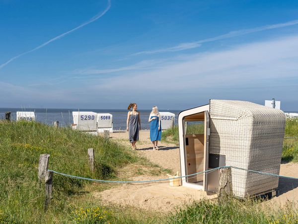 Zwei Frauen gehen, in Handtücher gekleidet zur geöffneten Strandkorbsauna.