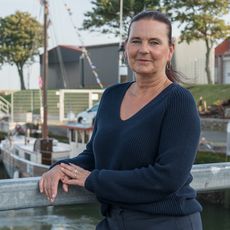 Eine Frau steht an einem Geländer. Hinter ihr ist Wasser und ein Bootsanleger.