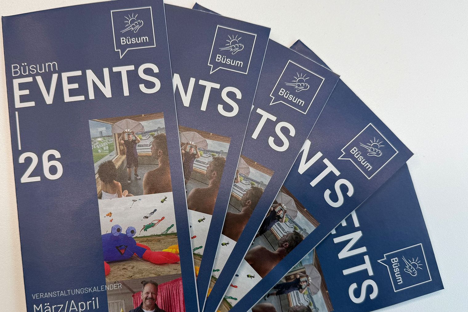 Mehrere Flyer in blauer Farbe mit der Aufschrift Events 26 März/April auf einem Tisch liegend.
