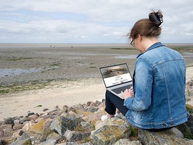 Frau sitzt mit Laptop am Büsumer Strand an der Steinkante und schaut sich Büsums neuen Internetauftritt an.