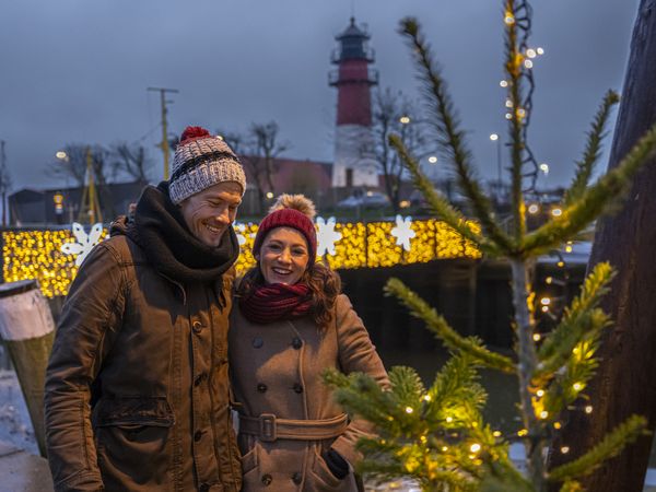 Paar geht Arm in Arm vor dem Leuchtturm am Museumshafen in der Adventszeit spazieren.