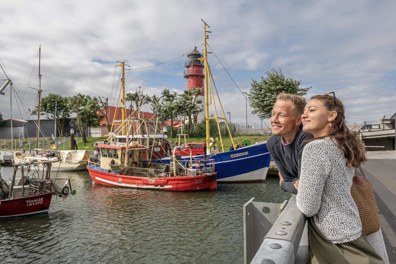 Ein junges Paar steht auf einer Brücke am Hafen. Im Hintergrund sind ein blauer und ein roter Kutter und ein Leuchtturm.