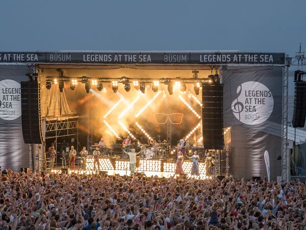 Eine hell erleuchtete Bühne mit Künstlern und Künstlerinnen. Davor ein großes Publikum.