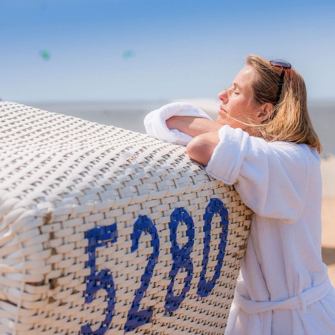 Eine Frau im Bademantel lehnt sich an einen Strandkorb und genießt die Sonne.