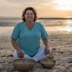 Eine Person am Strand. Sie sitzt im Schneidersitz am Strand. Vor ihr stehen zwei Klangschalen. Im Hintergrund ist das Wattenmeer und der Sonnenuntergang zu sehen.