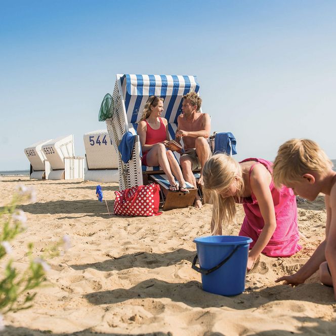 Zwei Erwachsene sitzen in einem Strandkorb, während zwei Kinder vor ihnen im Sand buddeln.