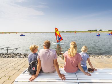 Familie sitzt auf einer Bank an der Familienlagune und schaut den Surfern zu. Im Hintergrund sieht man die Watt'n Insel und die Nordsee.