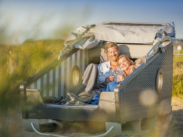 Mann und Frau liegen Arm in Arm in einem Schlafstrandkorb zwischen den Dünen. Die Sonne geht unter.