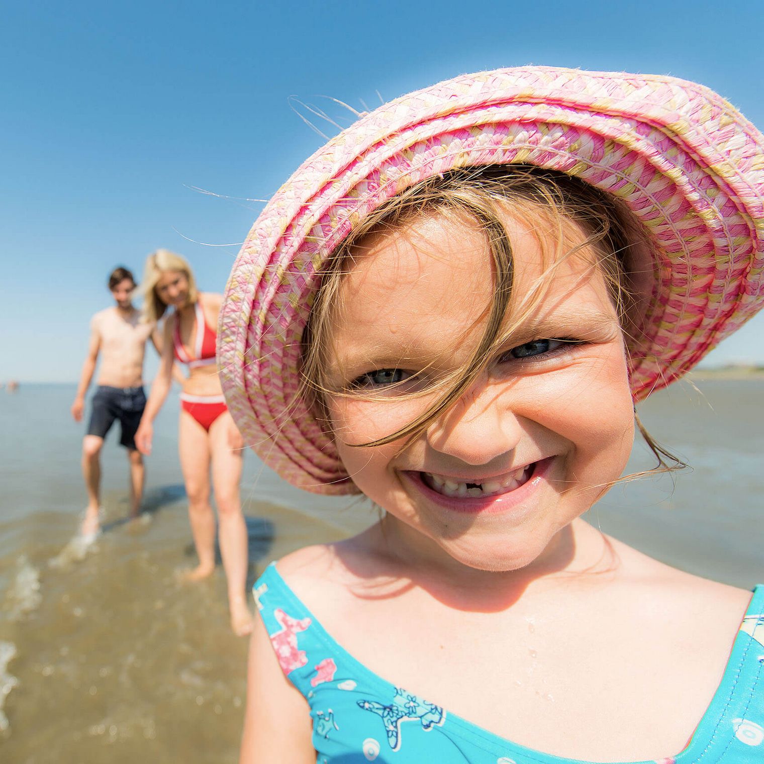 Ein kleines Mädchen mit Sonnenhut am Strand lacht in die Kamera.