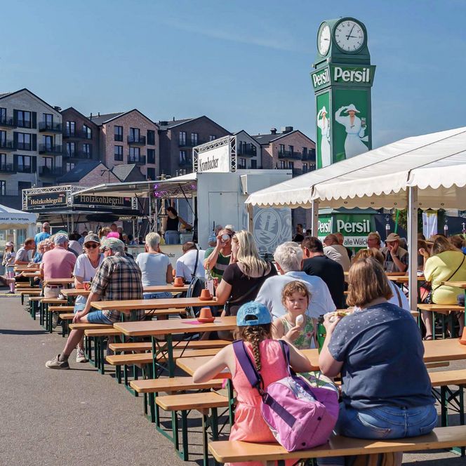 Personen sitzen auf Bänken in der Sonne auf dem Büsumer Winzerfest am Ankerplatz.