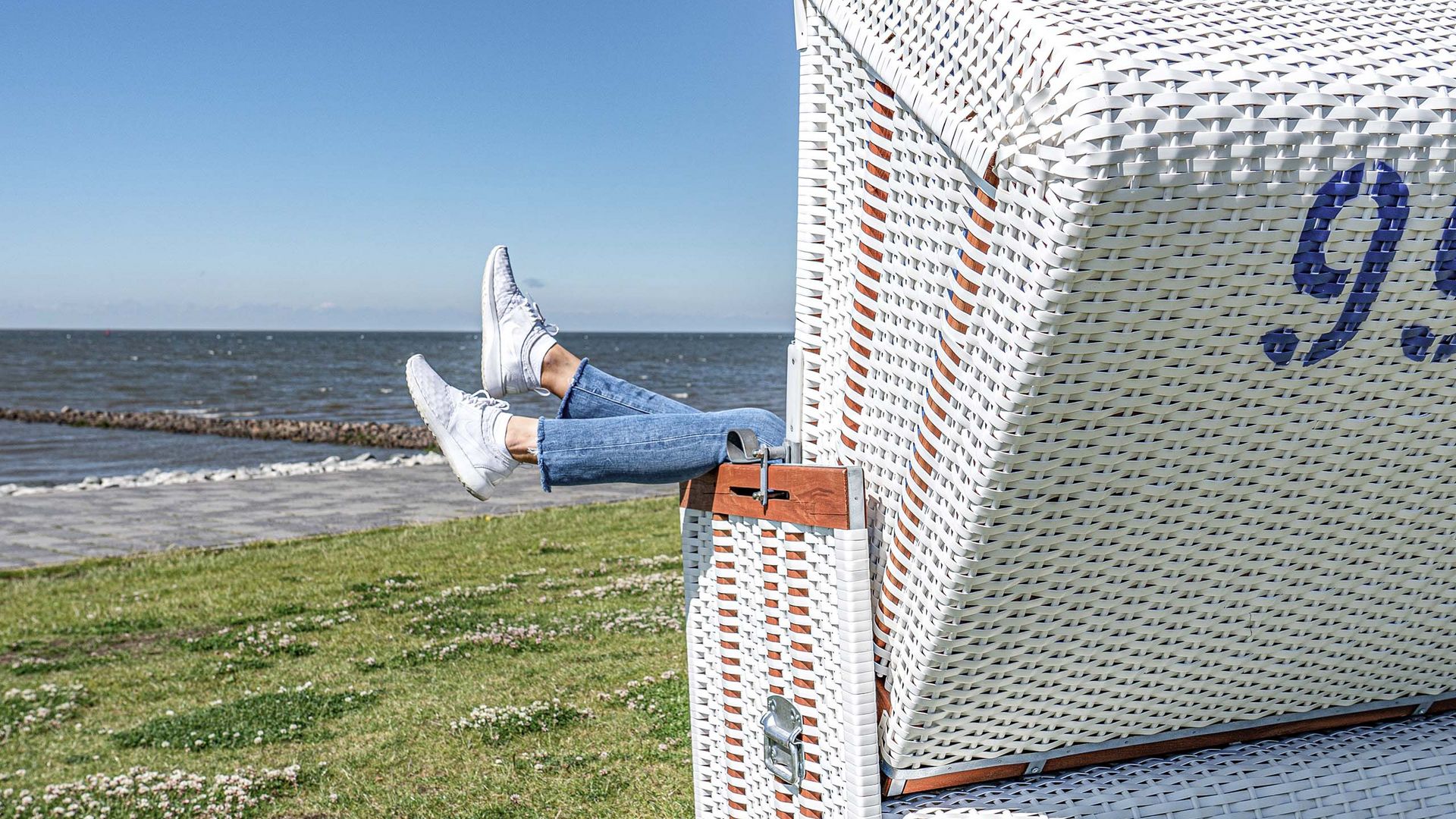 Eine Person hält ihre Füße aus einem Strandkorb raus und im Hintergrund ist die Nordsee zu sehen.