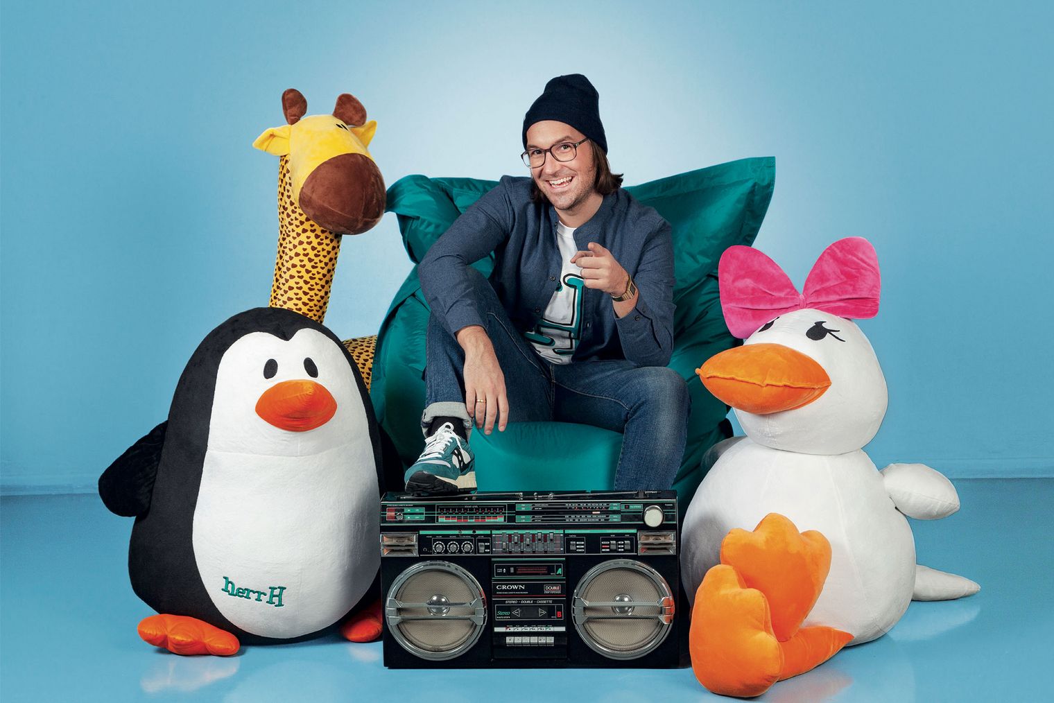 Ein Mann mit Mütze in einem Sitzsack. Davor ein Radio, links und rechts Plüschtiere, eine Giraffe, ein Pinguin und eine Ente.