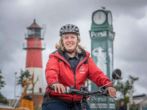 Die Büsumer Gästelotsin auf einem Fahrrad vor dem Büsumer Leuchtturm und der Persil-Uhr am Ankerplatz.