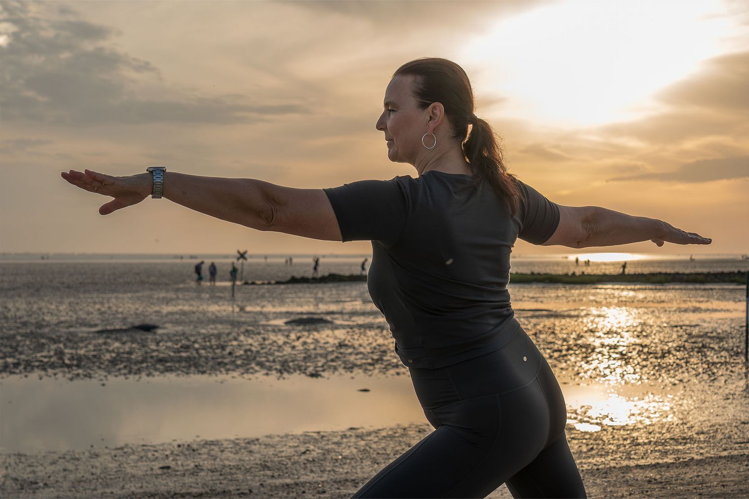 Eine Person am Strand in der Krieger Pose aus dem Yoga. Im Hintergrund ist das Wattenmeer und der Sonnenuntergang zu sehen.