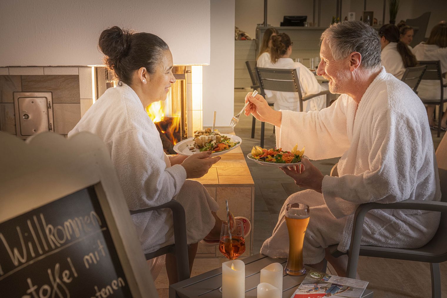 Ein Paar, sitzend vor einem Kamin. Sie halten jeweils einen Teller mit Essen in der Hand. Sie tragen beide einen Bademantel Vor ihnen stehen zwei Getränke.