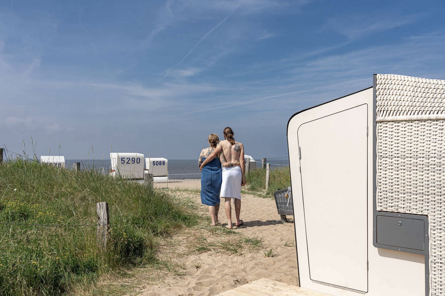 Ein Paar läuft von der Strandkorbsauna aus über den Strand in Richtung Nordsee.